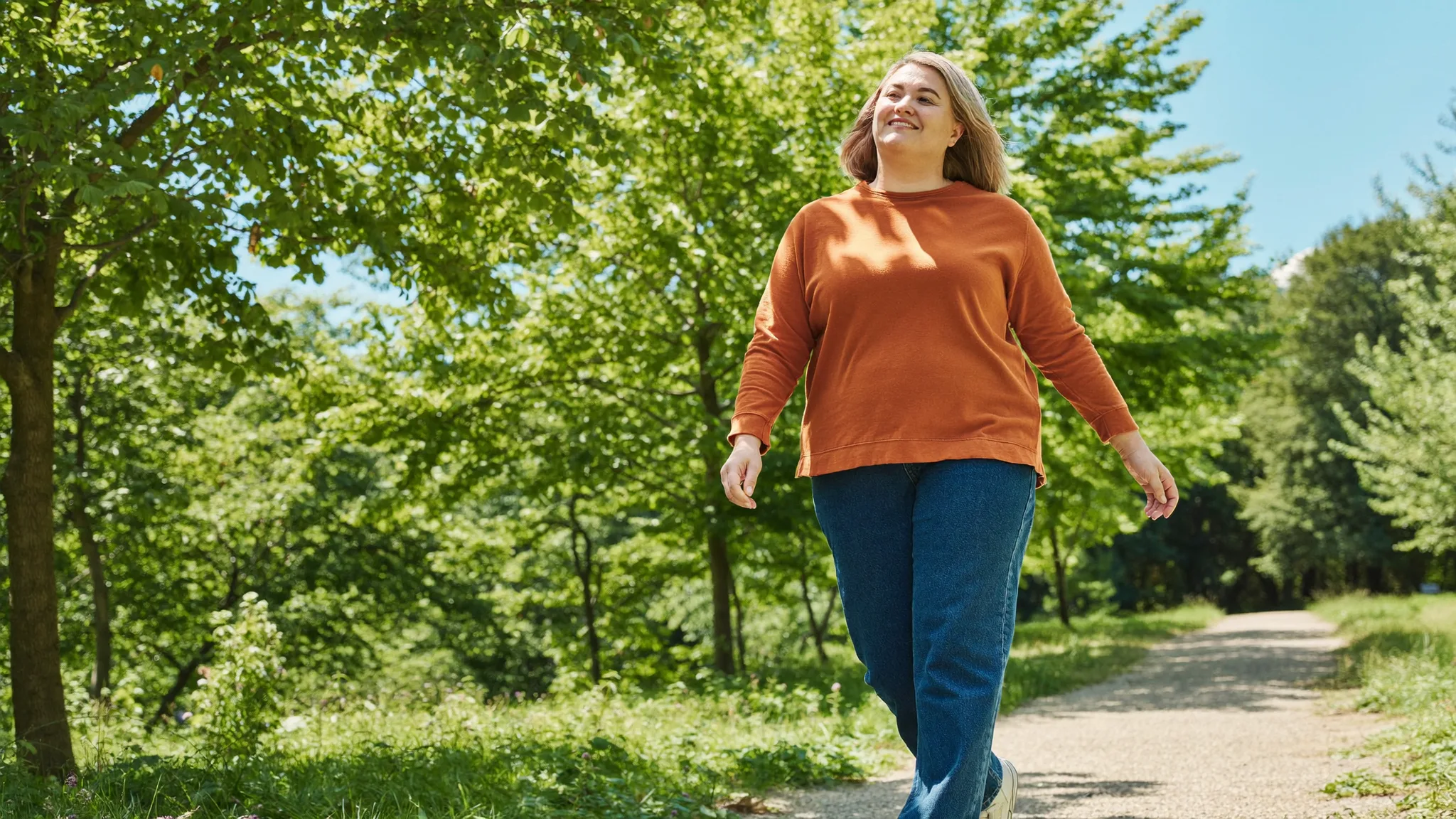 A woman in a rust-coloured sweatshirt walking along a leafy park path