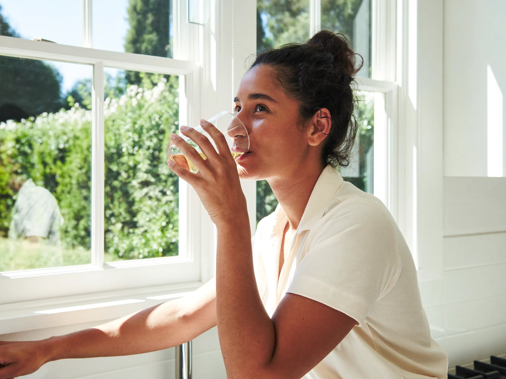 A woman drinking a glass of water by a sunny kitchen window