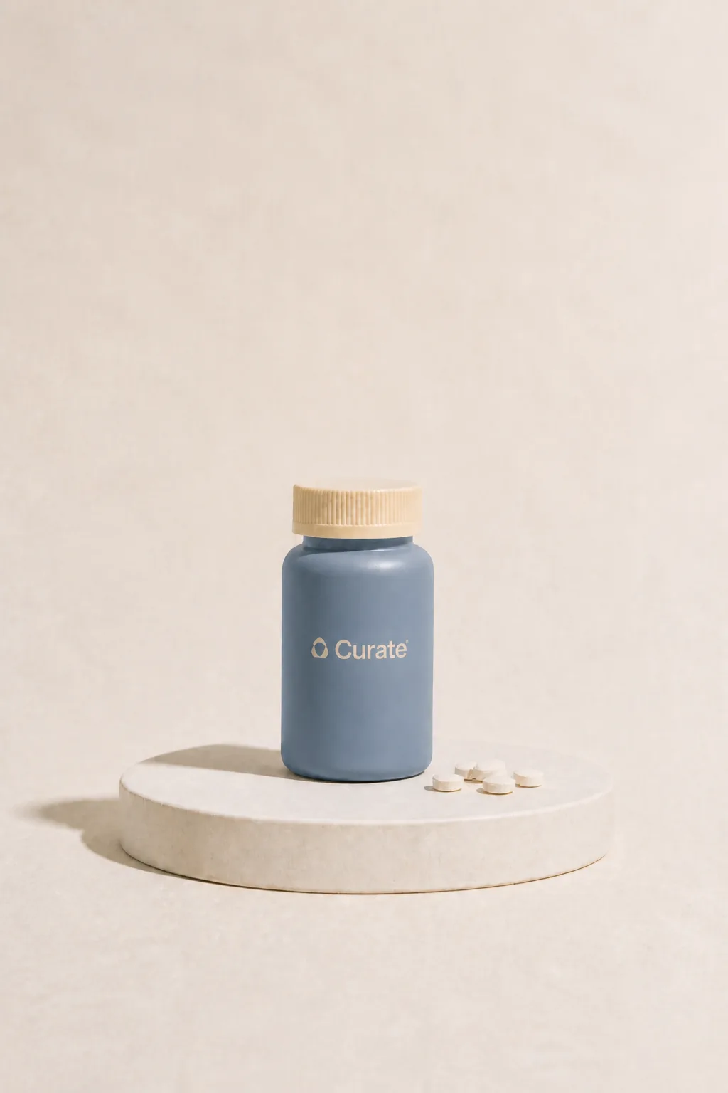 A slate-blue Curate pill bottle with loose white tablets on a studio plinth