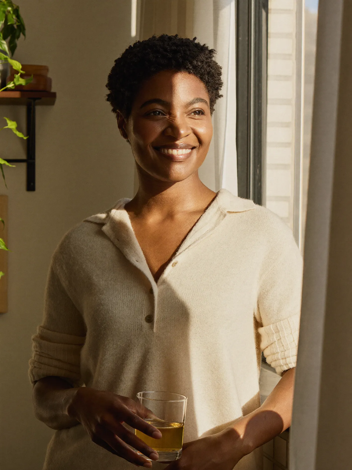 A woman at a bright kitchen window in soft morning light, holding a glass of water