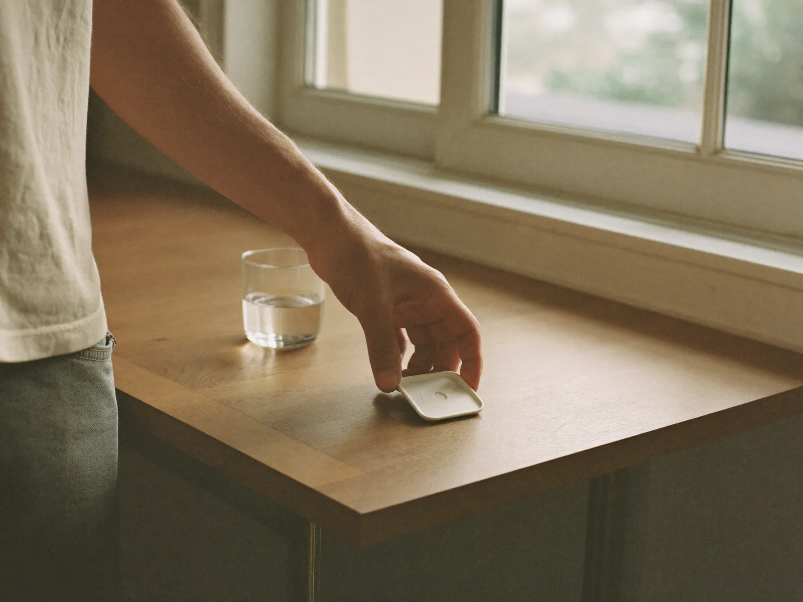 A hand picking up a small tablet beside a glass of water in morning light
