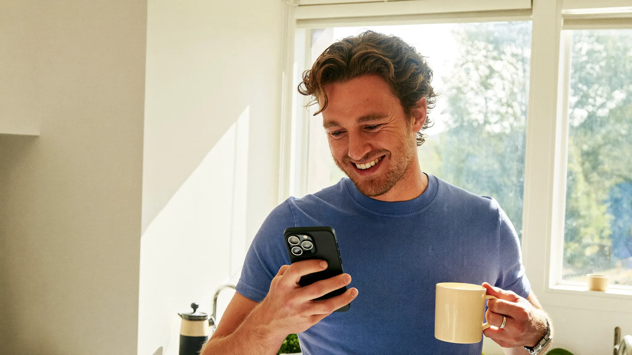 A man in a sunlit kitchen smiling at a message on his phone